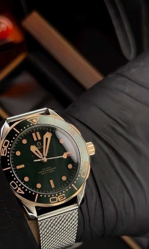 Silver wristwatch with a green face and mesh strap on a black sleeve.
