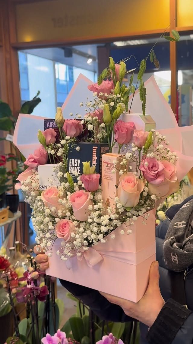 Pink gift box filled with roses, baby's breath, and beauty products held by hands.