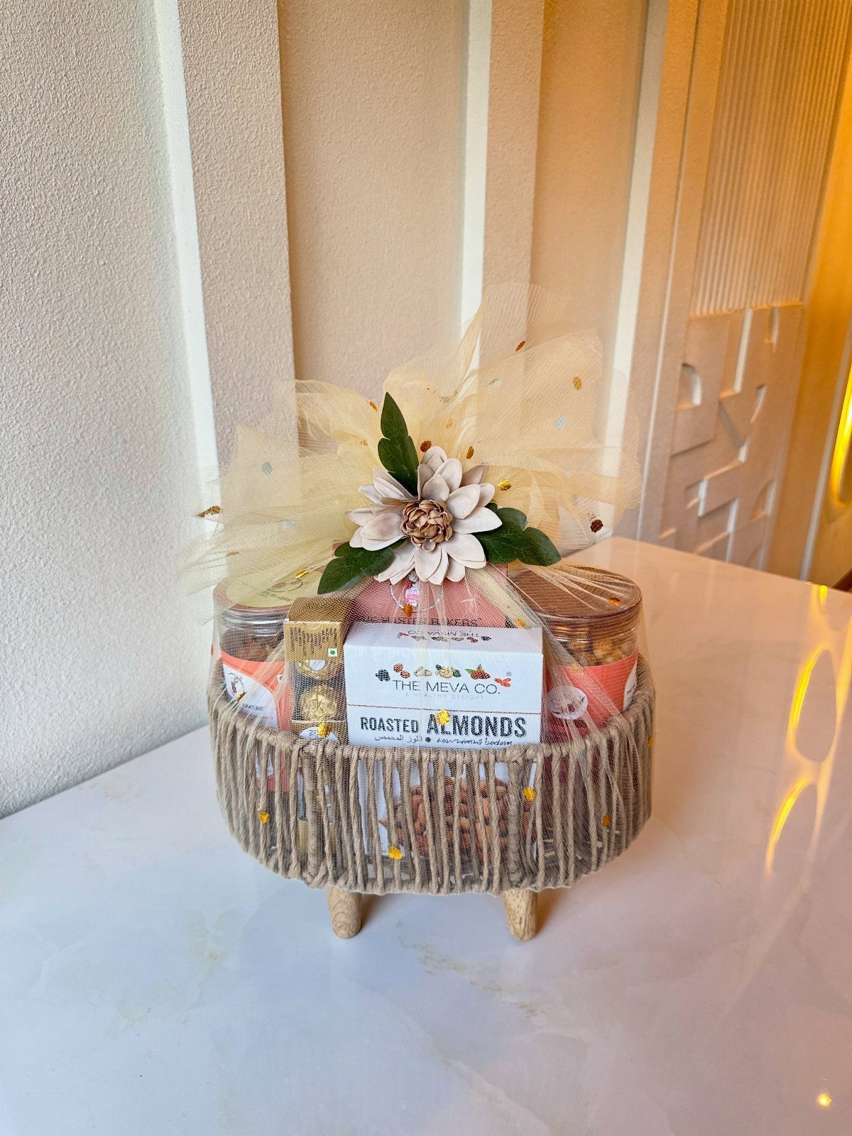Woven rope gift basket filled with gourmet snacks and decorated with a white flower.