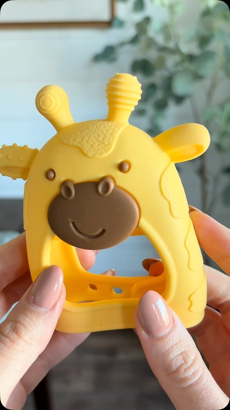Yellow silicone giraffe baby teether held by two hands in a blurred room.