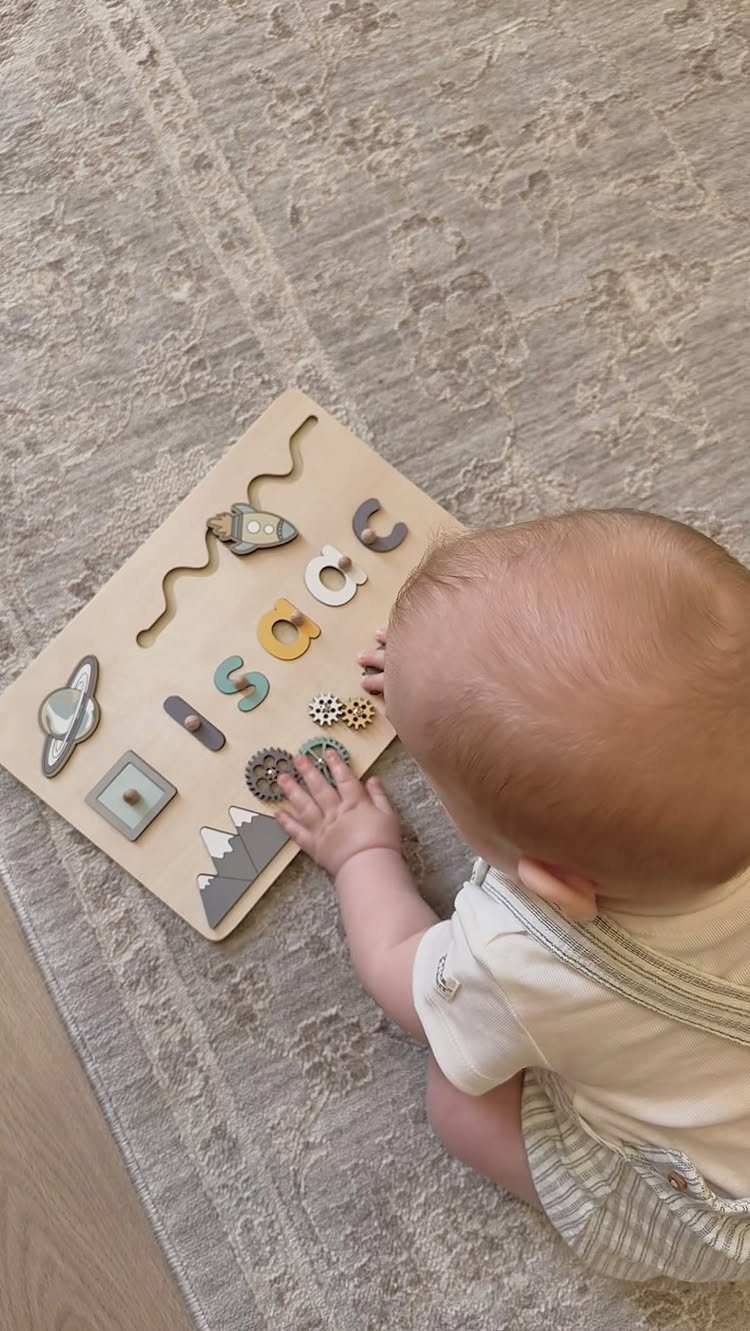 Baby playing with a wooden personalized name puzzle on a grey rug.