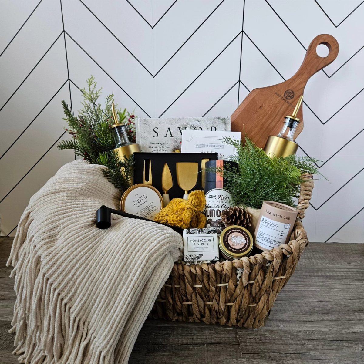Woven gift basket with a beige throw, gold utensils, and autumn-themed gourmet treats.