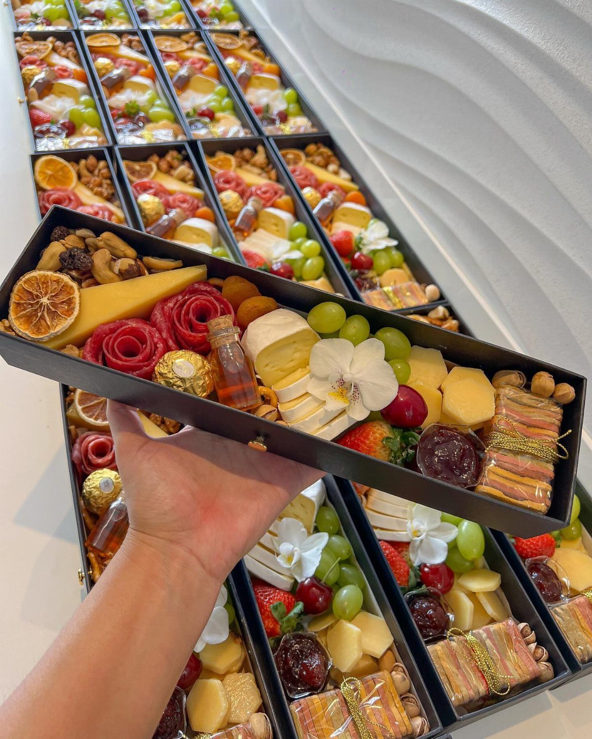 Gourmet food gift box filled with cheeses, fruits, and nuts held by a hand.