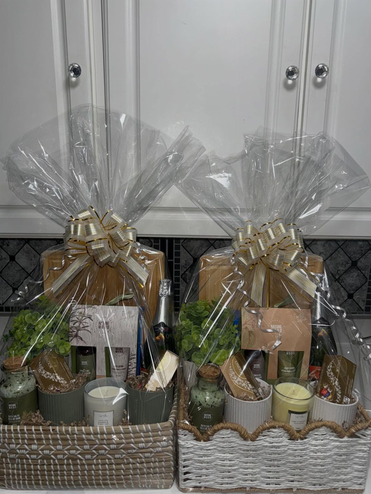 Two wrapped gift baskets containing cutting boards, candles, and plants for a host.