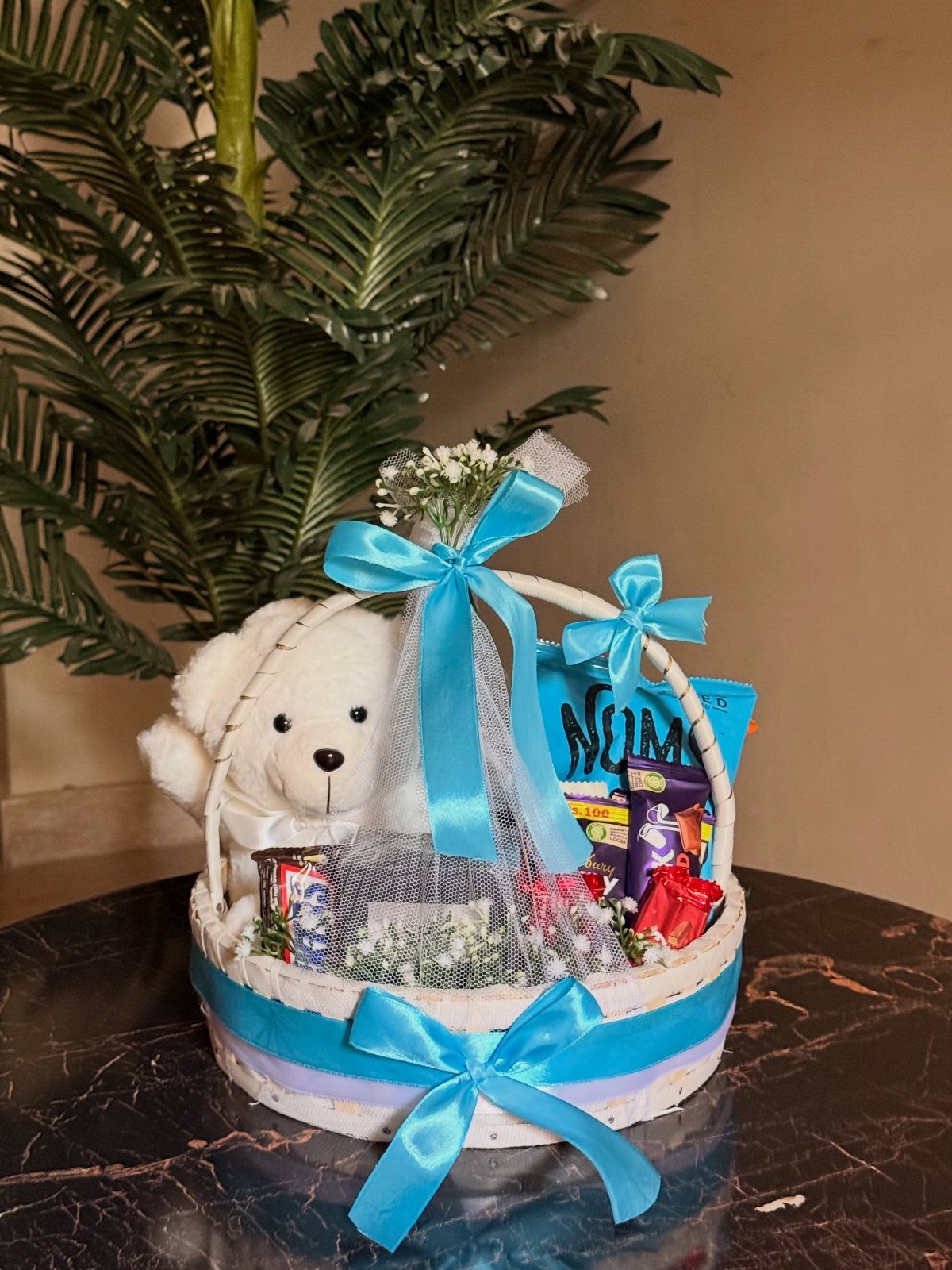 White wicker gift basket with a teddy bear, chocolates, and blue satin ribbons.