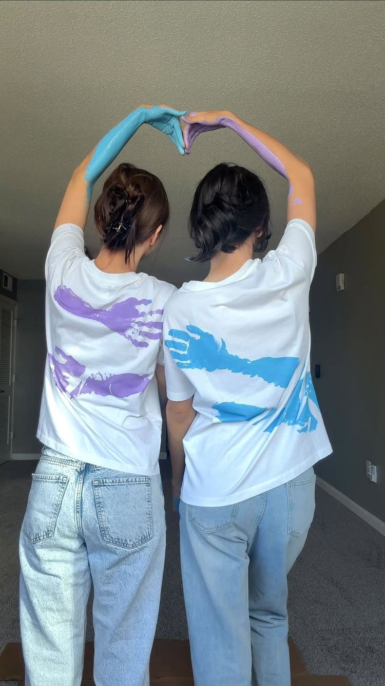 Two women wearing white t-shirts with colorful handprints forming a heart with hands.