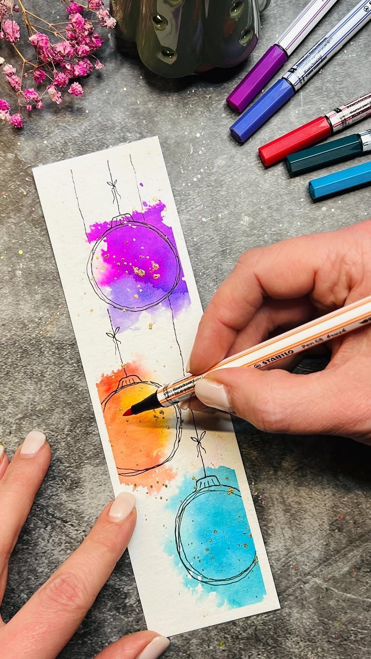 Hands painting colorful watercolor Christmas ornaments on a strip of white paper.