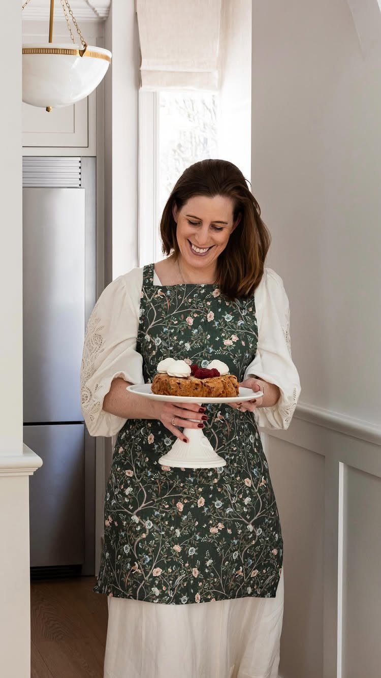 Woman wearing a dark green floral apron holding a cake on a stand.