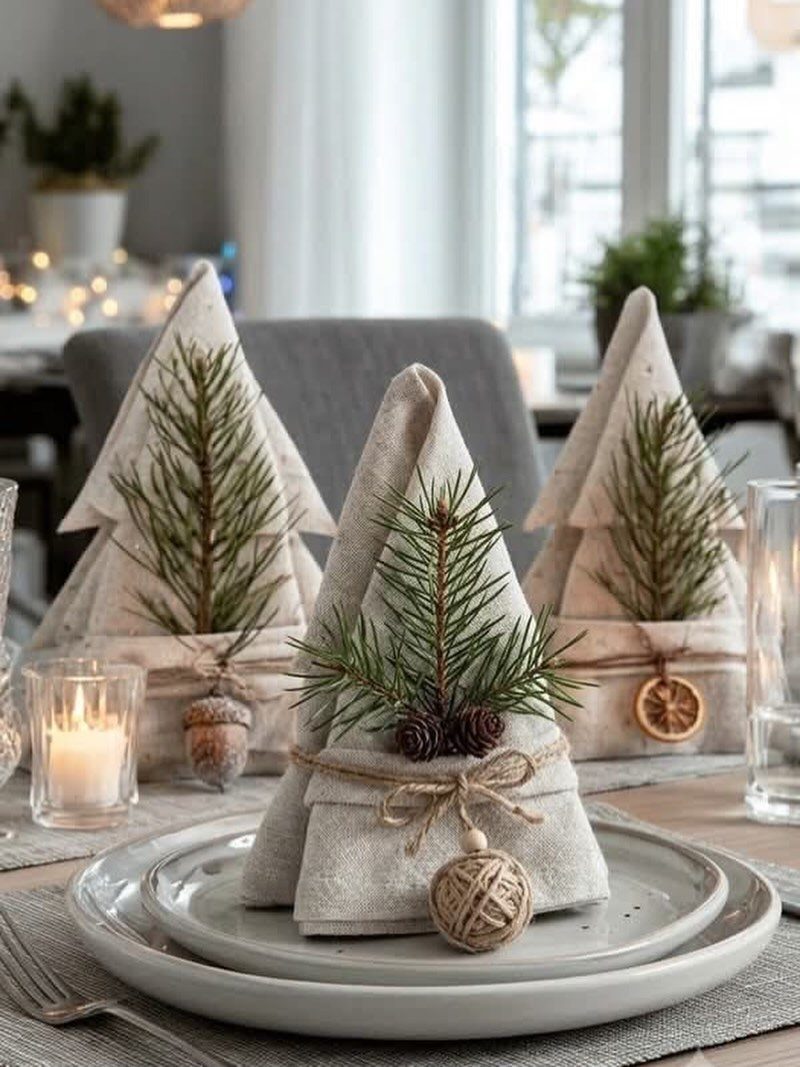 Beige linen napkins folded as Christmas trees with pine sprigs on white plates.