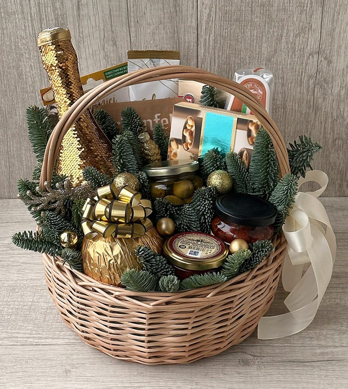 Woven gift basket with gold bottle, gourmet jars, and pine branches for Christmas.
