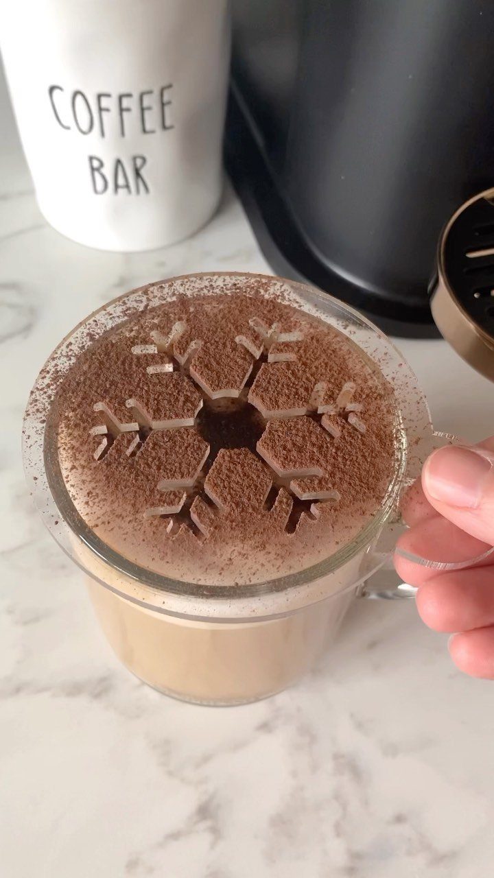 15 Coffee Drinker Gifts: Candles, Mugs, Roasters and More 5 Hand lifting a snowflake stencil from a coffee drink topped with cocoa powder