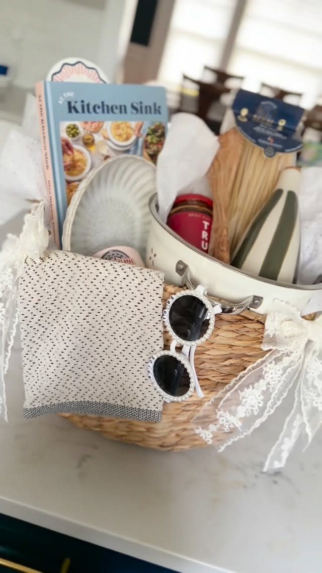 Wicker gift basket containing a cookbook, pasta, and kitchen accessories on a counter.