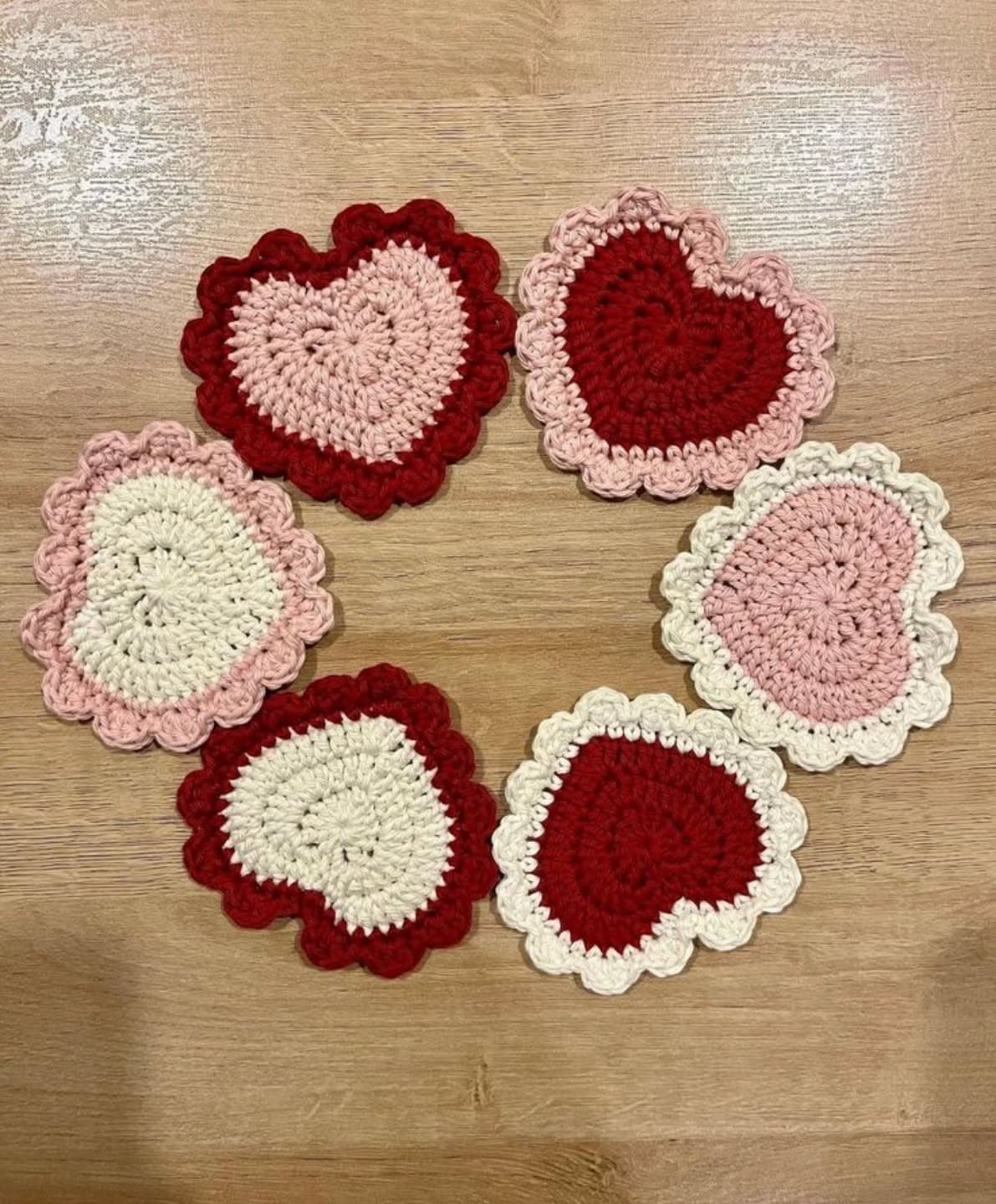 Six heart-shaped crochet coasters in red, pink, and cream on a wooden table.