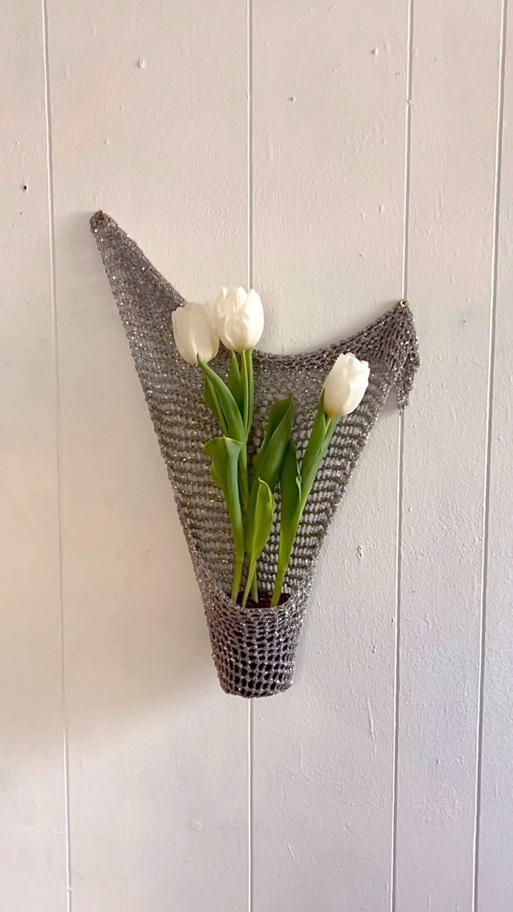 Grey crochet wall pocket holding three white tulips against a white wooden wall.