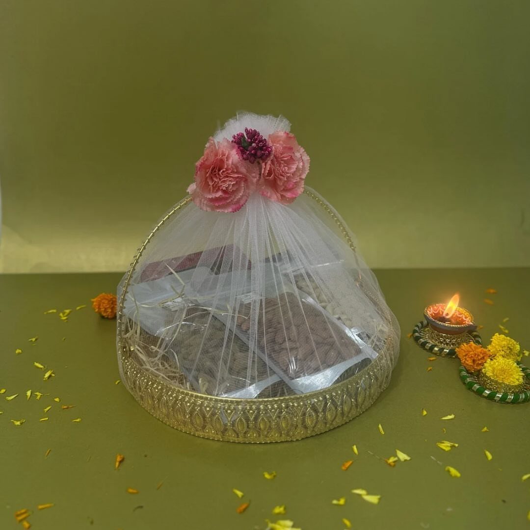 Diwali Gift Hampers: 8 Ideas for Sweets, Dry Fruit & More 6 Decorative gold tray with sweets under white tulle, pink flowers, and lit diya.