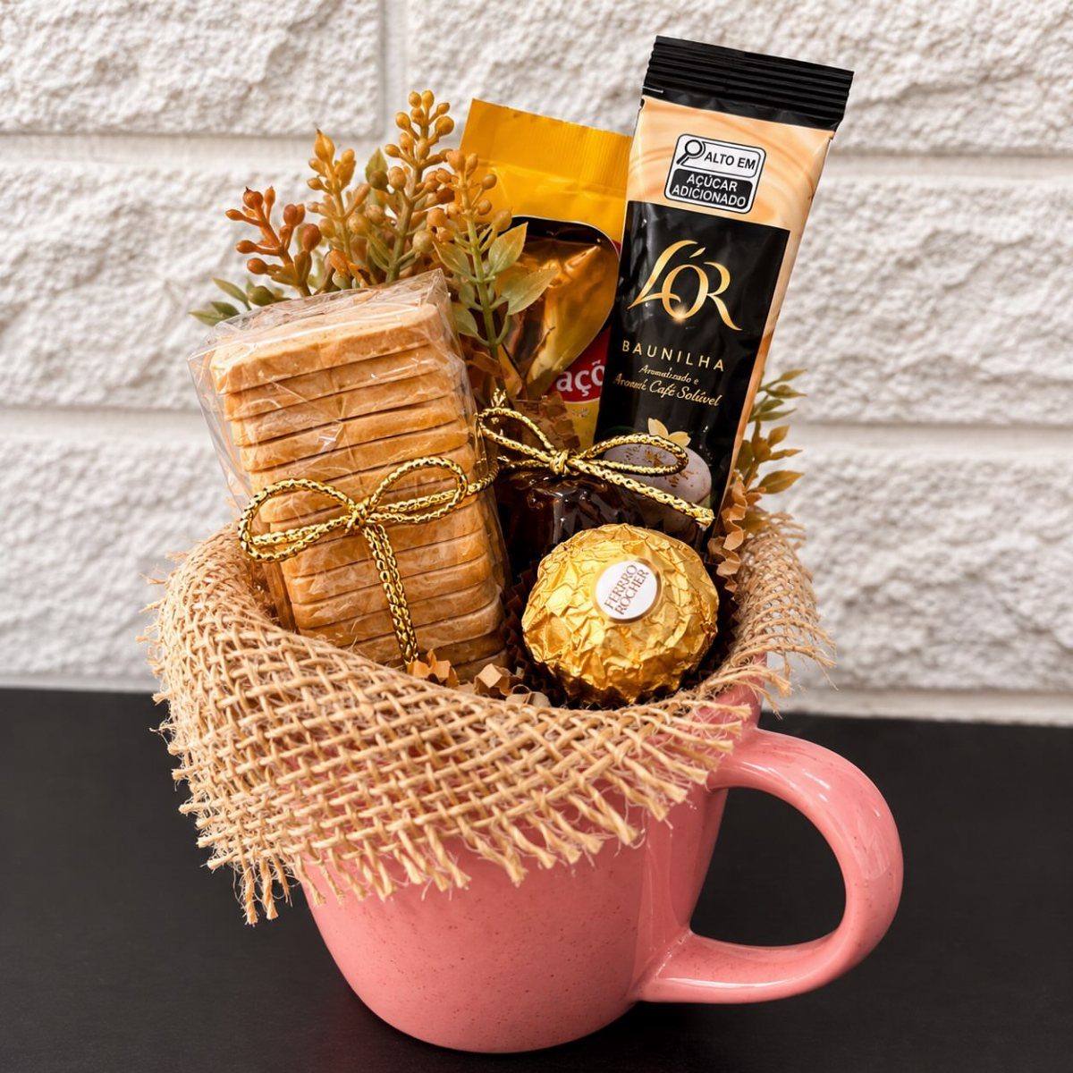 Pink mug gift basket with coffee pods, biscuits, and chocolate wrapped in burlap.