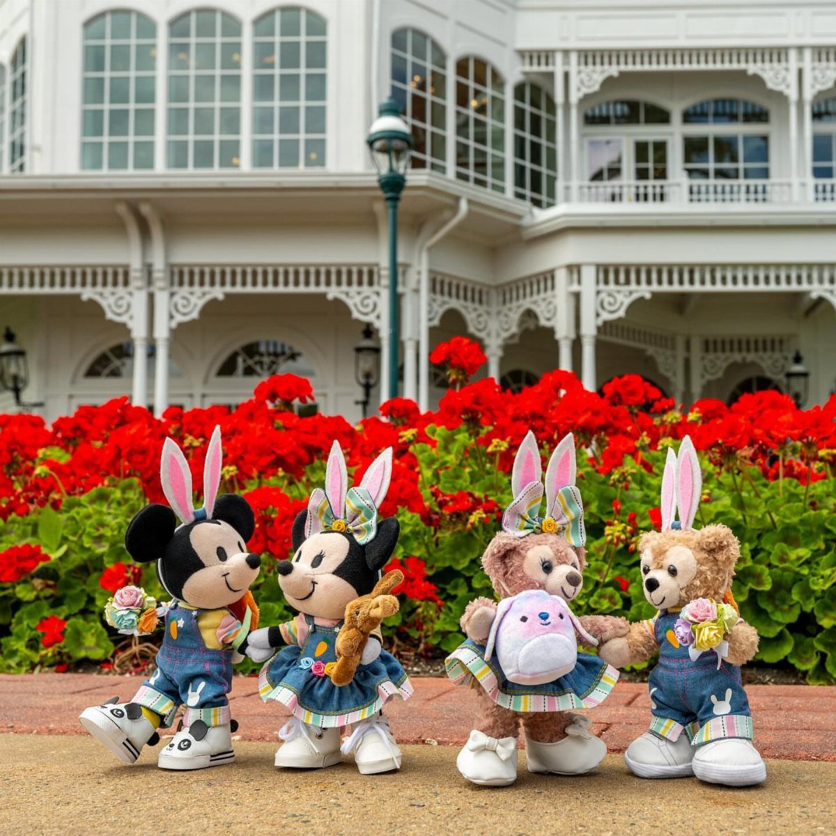 Four plush characters wearing bunny ears and denim outfits in front of red flowers.