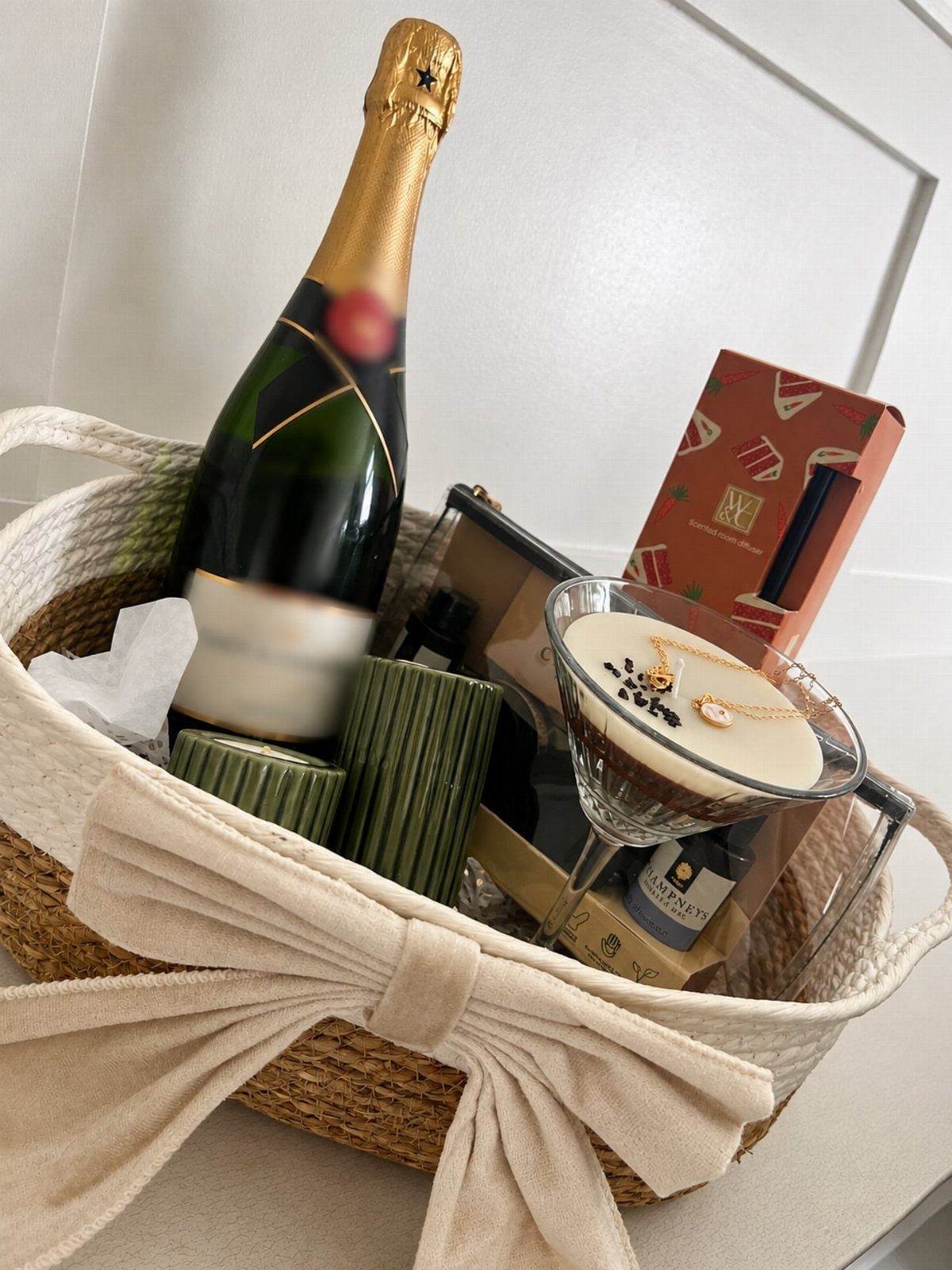 Gift basket with champagne, dessert, and treats tied with a large beige bow.