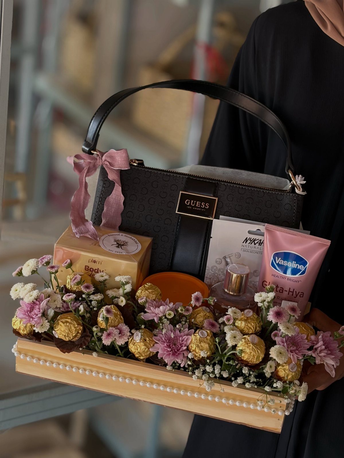 Gift tray with black Guess handbag, skincare products, chocolates, and pink flowers.