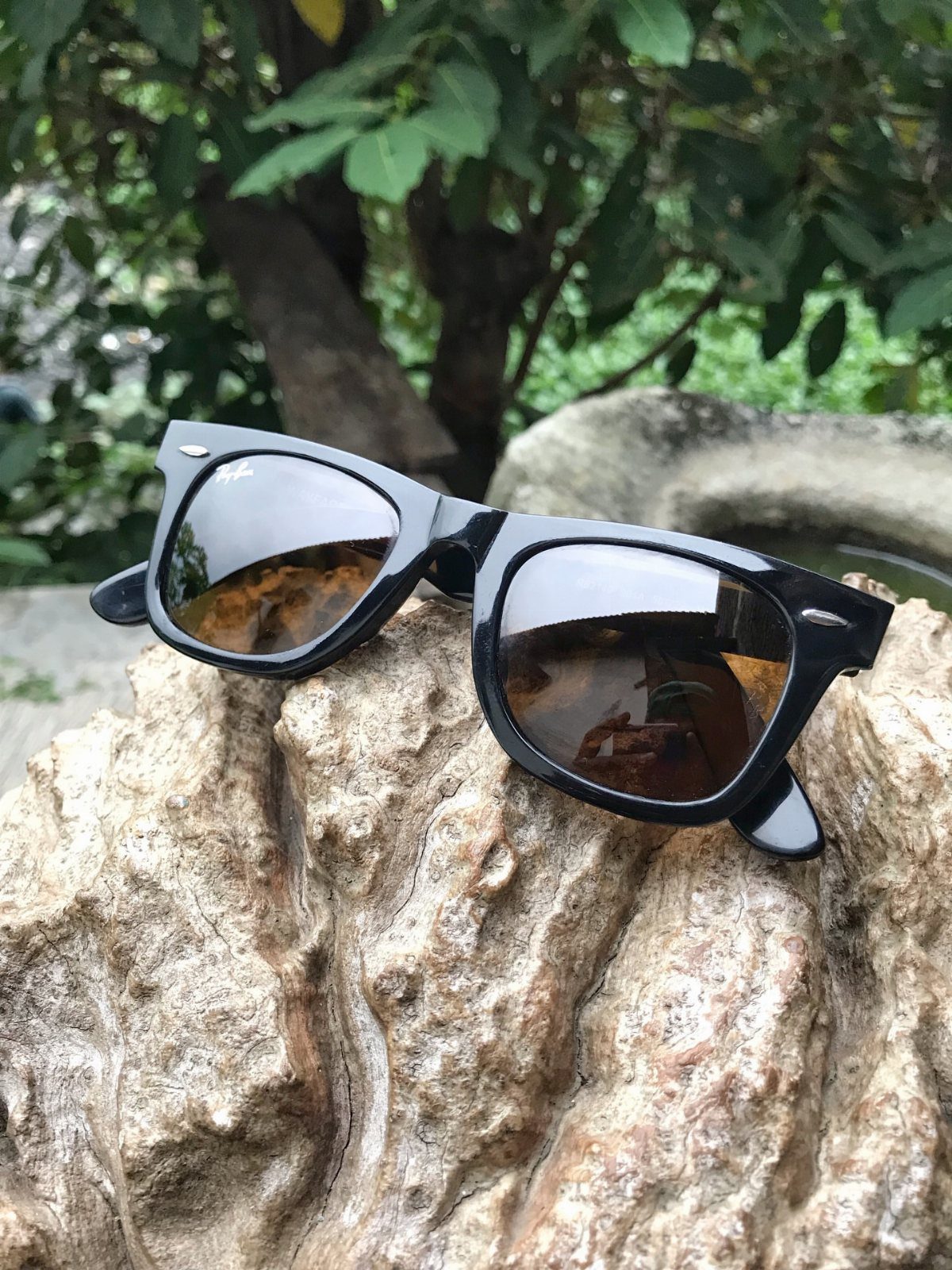 7 Father in Law Gift Ideas He'll Actually Use 6 Black square sunglasses resting on a textured light brown rock with green leaves behind.