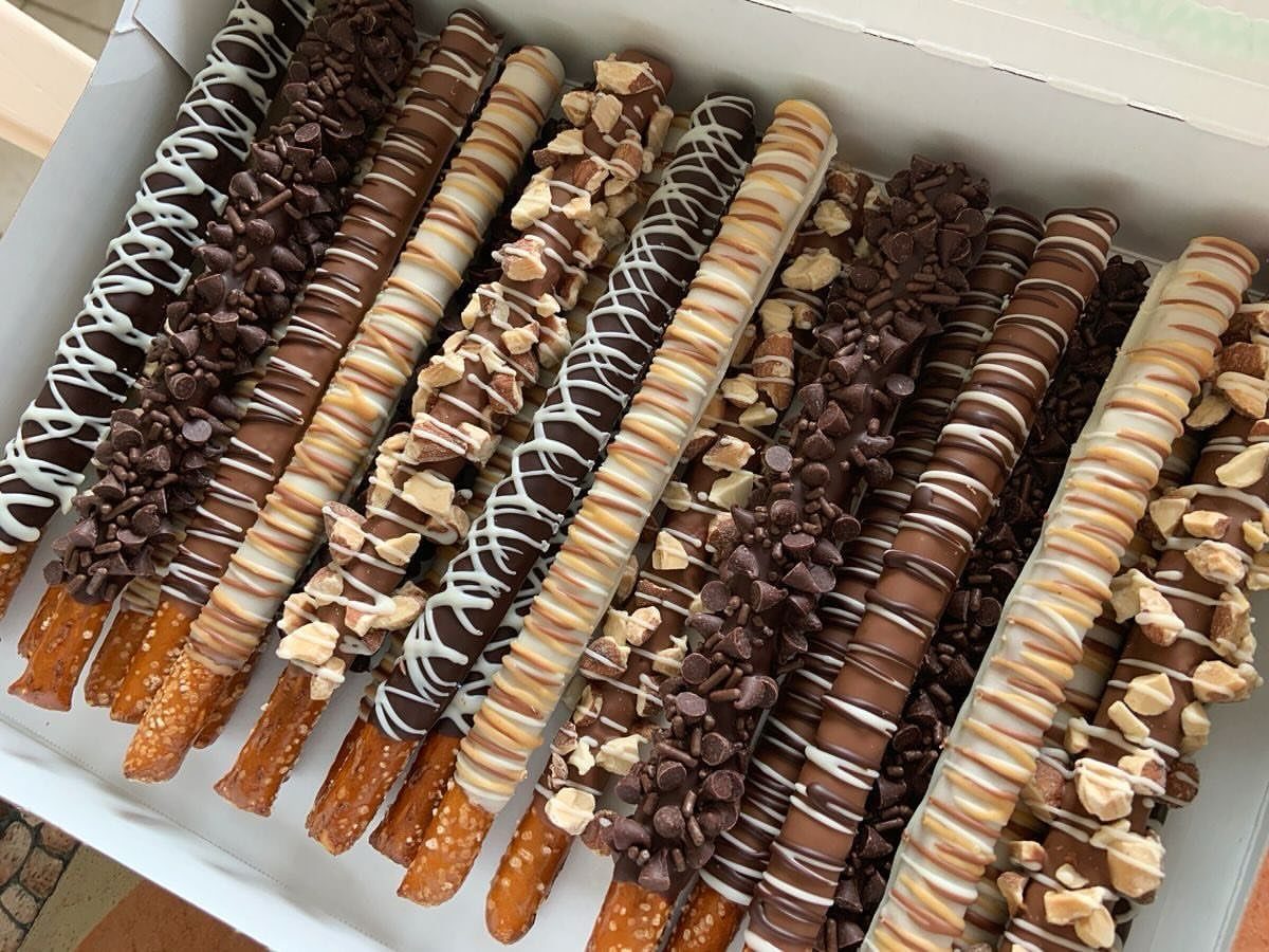 Chocolate-covered pretzel rods with various drizzles and nut toppings in a white box.