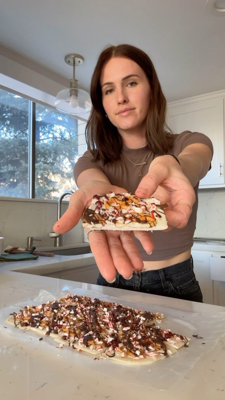 Woman holding a piece of peppermint chocolate bark in a bright kitchen.