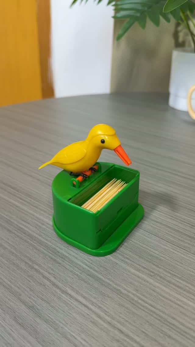 Yellow plastic bird perched on a green toothpick holder on a grey table.