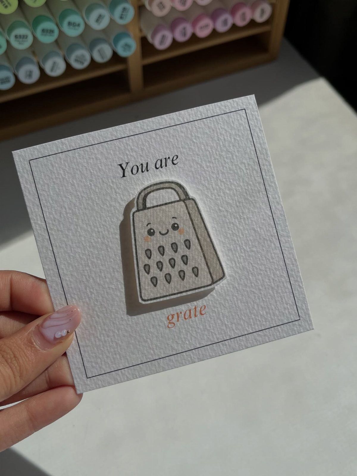 Hand holding a punny card with a cute cheese grater illustration and text.