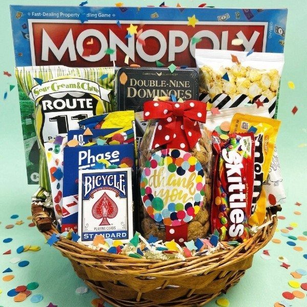 Wicker gift basket containing board games, playing cards, and various snacks with colorful confetti.