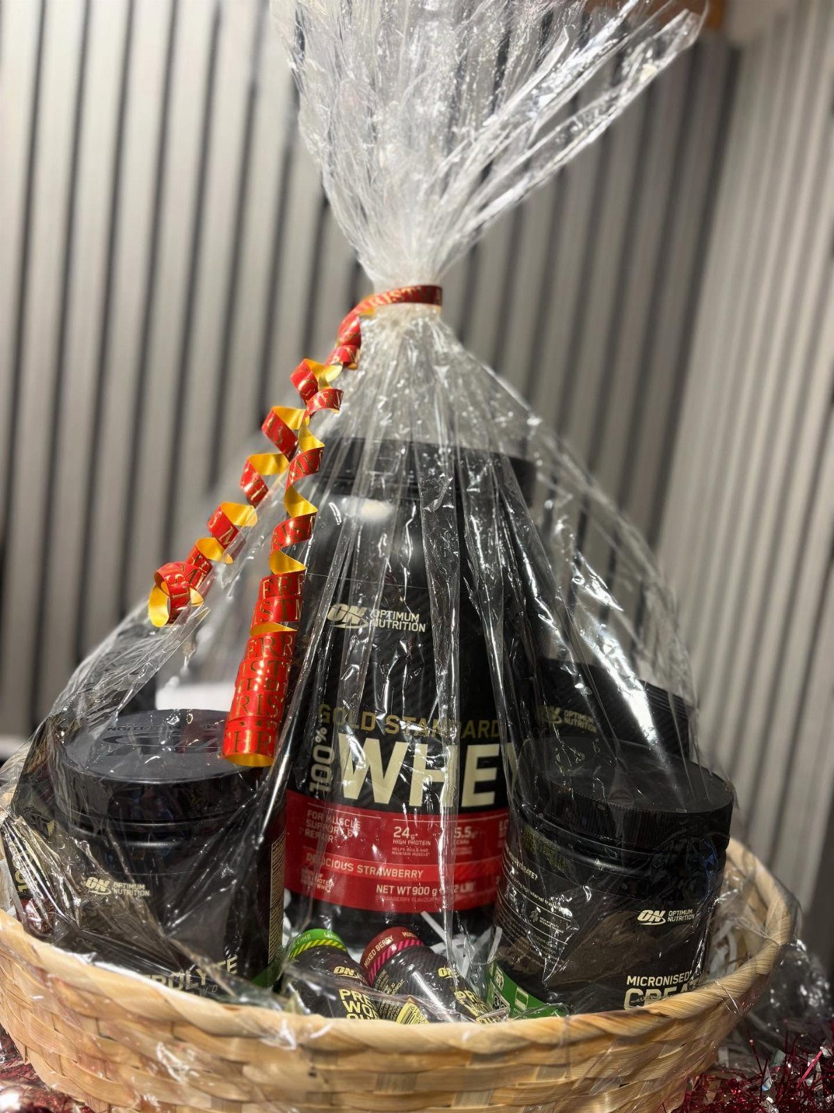 Wicker gift basket wrapped in cellophane containing protein powder and fitness supplements.