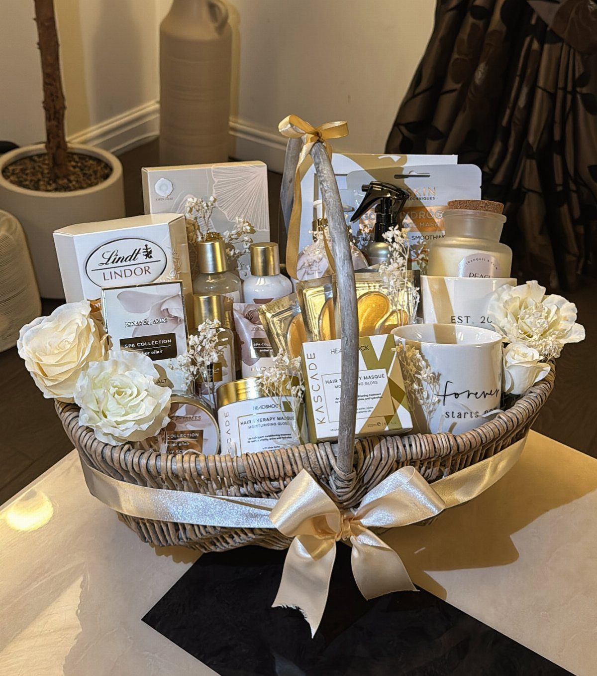 Wicker gift basket with skincare products, chocolates, and white roses tied with gold ribbon.