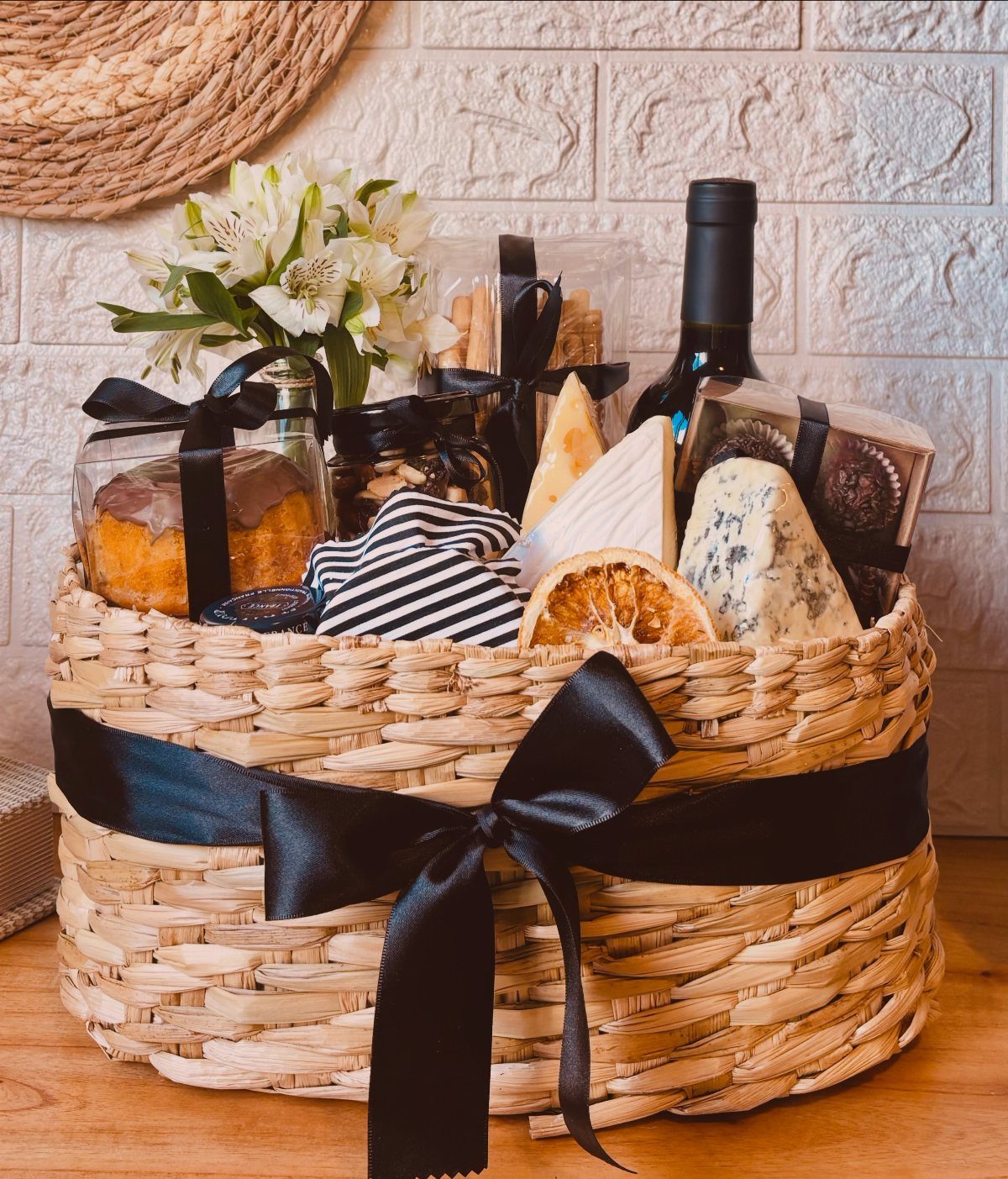 Woven gift basket with red wine, gourmet cheeses, cake, and white flowers.