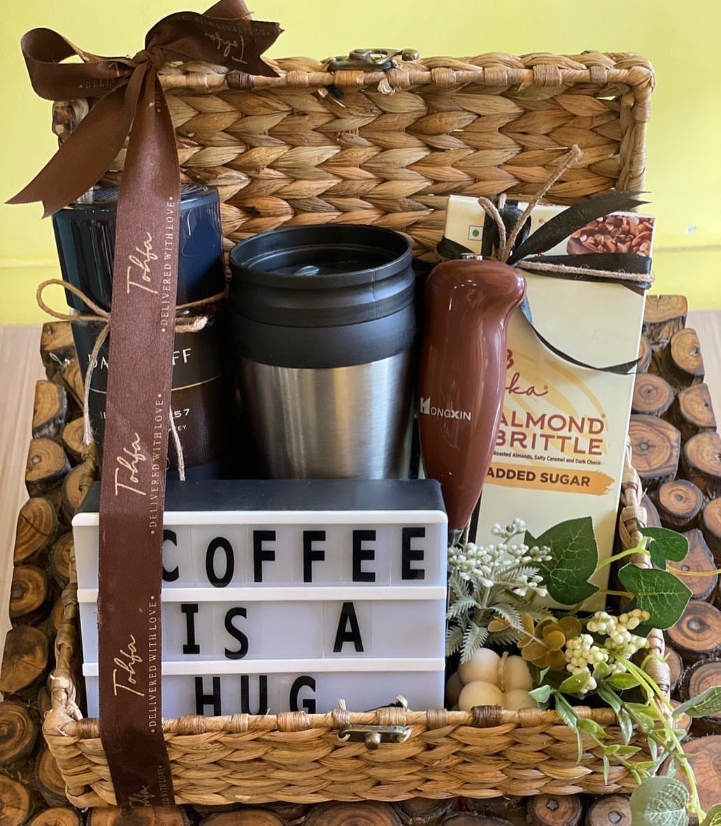12 Gift Ideas for the Office Your Coworkers Will Use 6 Wicker gift basket with travel mugs, almond brittle, and a coffee lightbox sign.