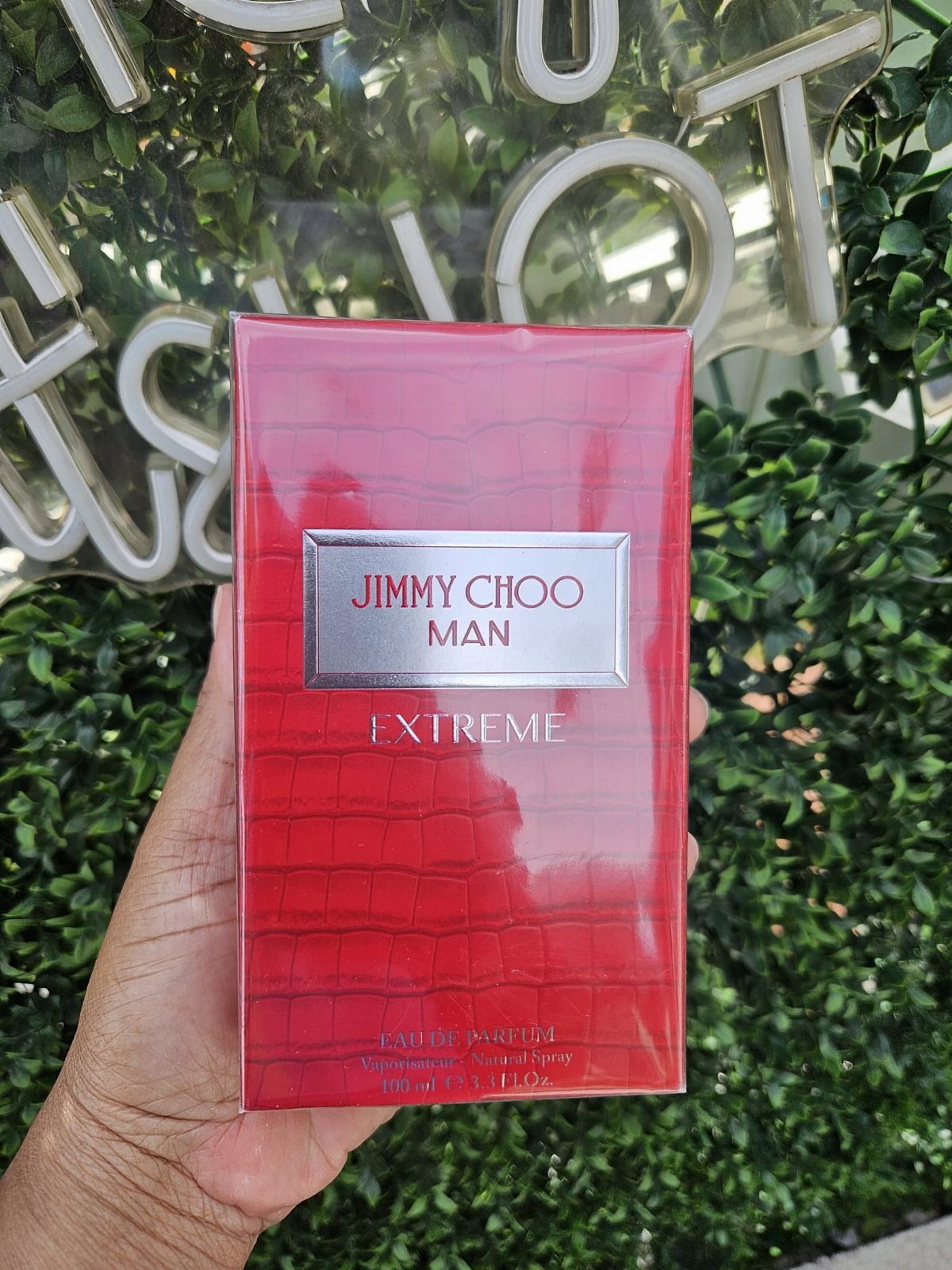 Red Jimmy Choo Man Extreme cologne box held against a green leaf background.