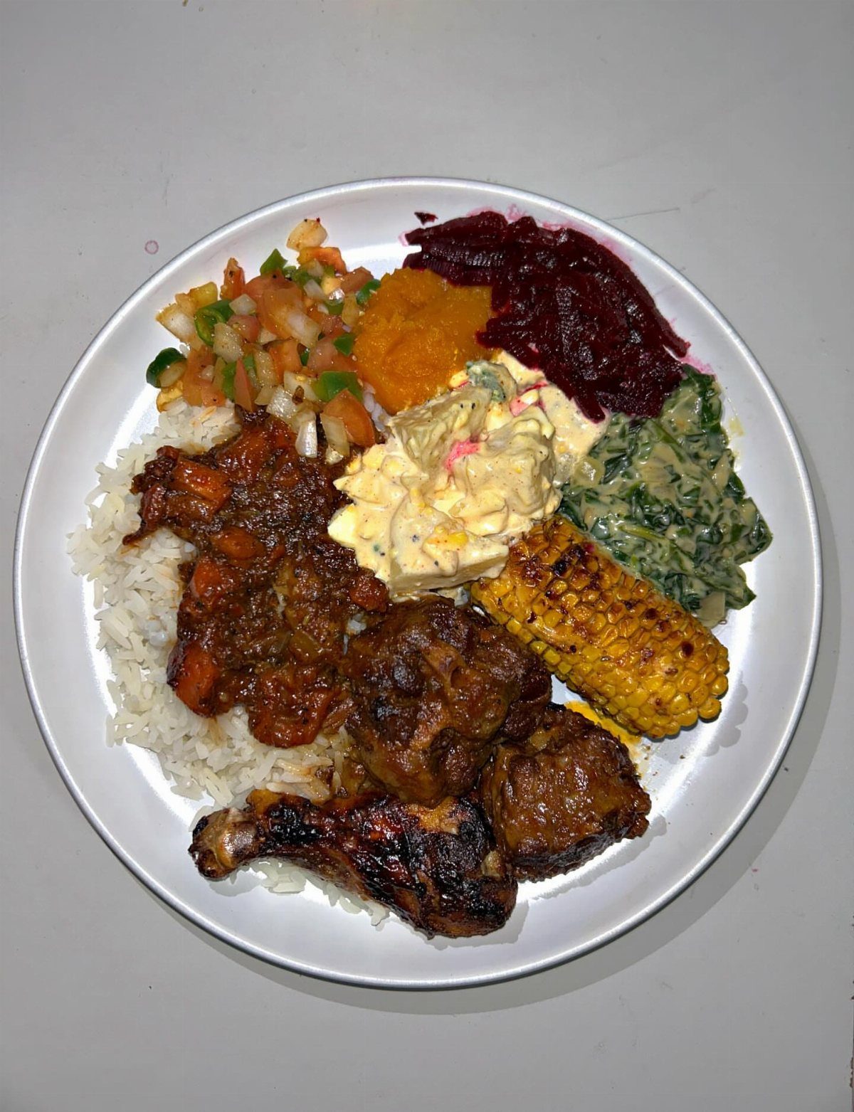 Platter of grilled chicken, corn, rice, and assorted vegetable sides on white plate.