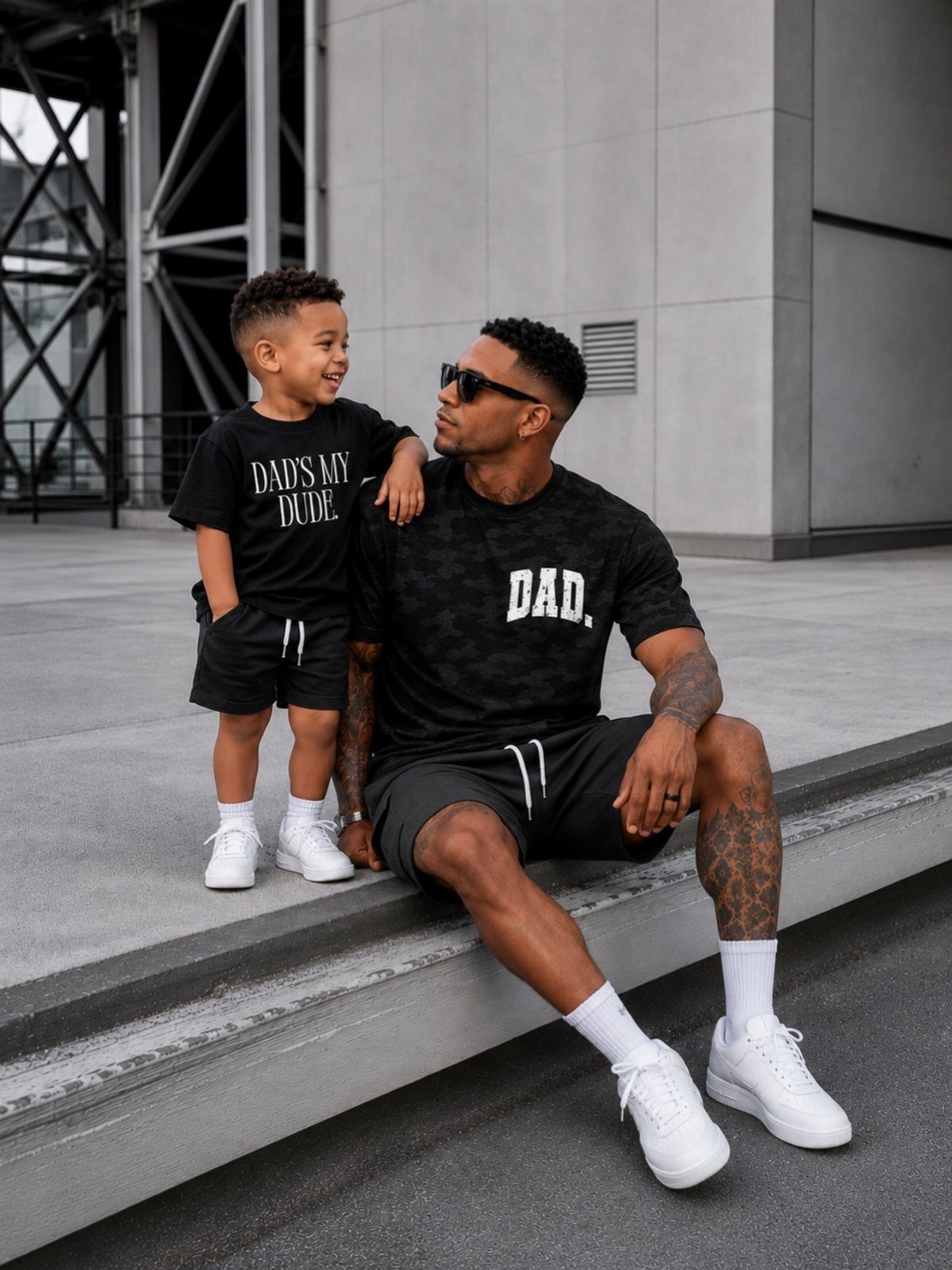 7 Gifts for Dad From Son That Feel Personal and Fun 3 Father and son wearing matching black t-shirts and shorts sitting outdoors.