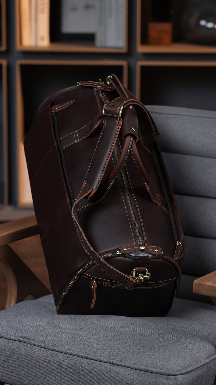 Dark brown leather duffel bag resting on a grey fabric armchair in a room.