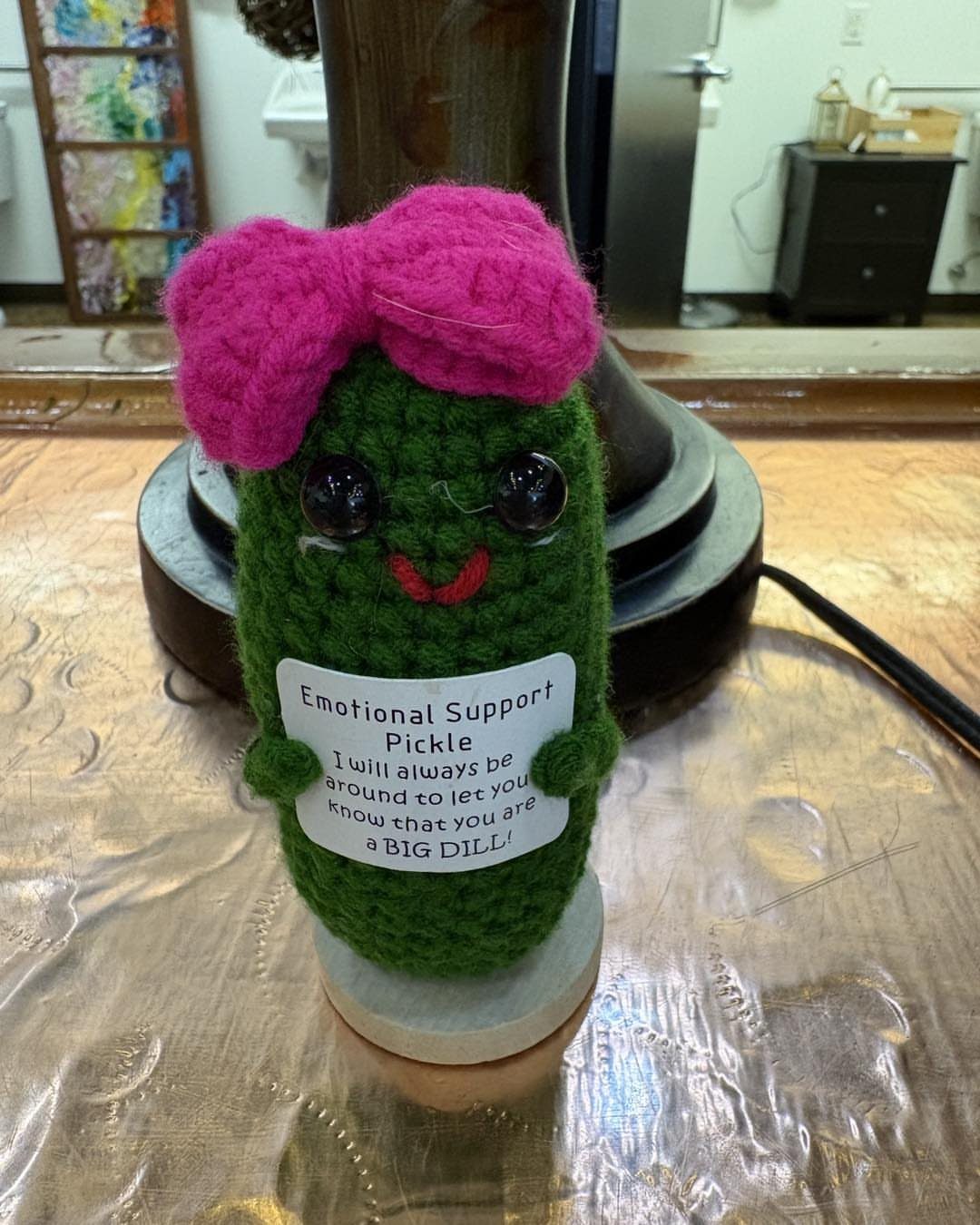Crocheted green pickle plush with a pink bow holding an emotional support card.