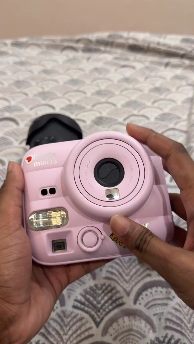 Hands holding a light pink instant camera over a patterned grey fabric background.