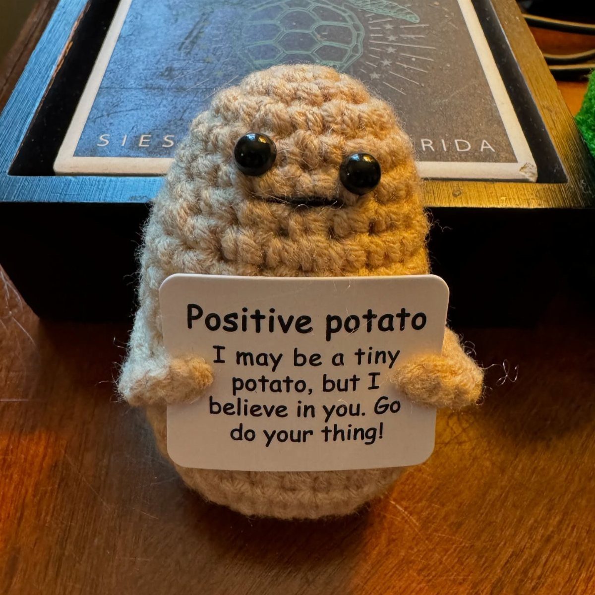 Crocheted potato plush holding a positive affirmation sign on a wooden table.