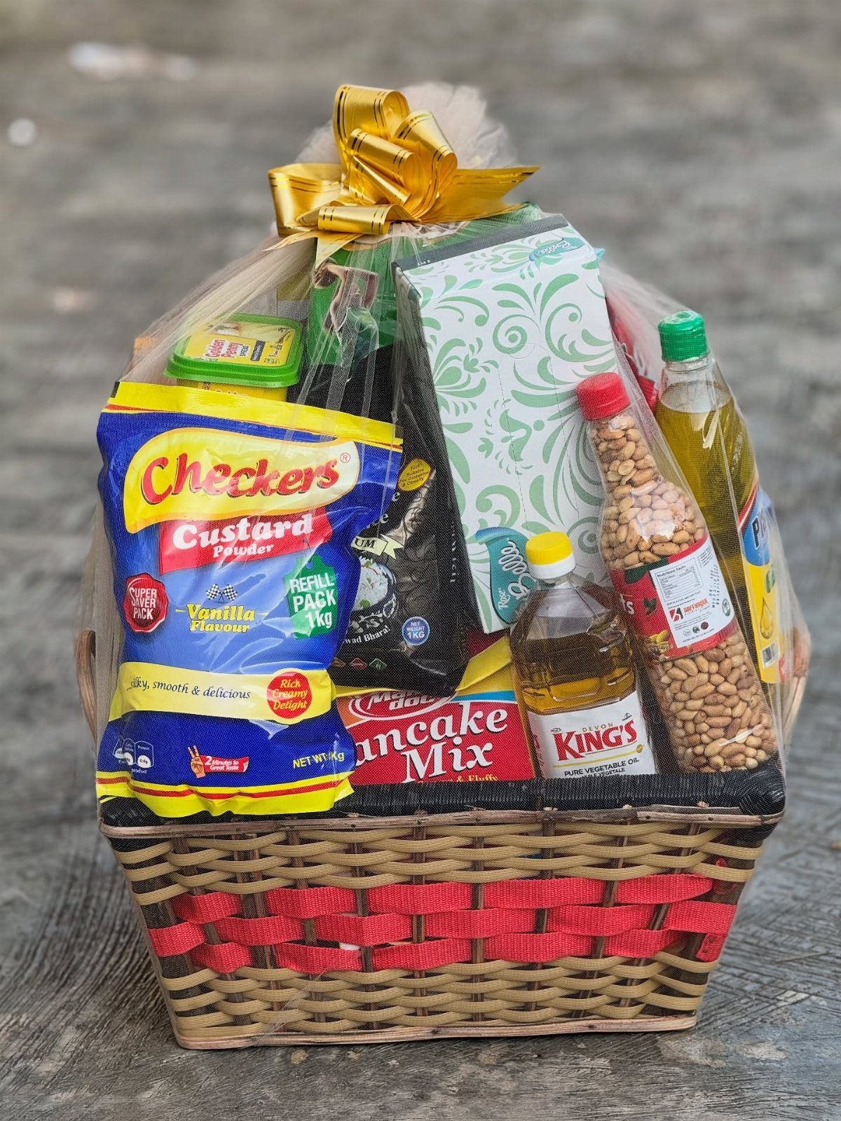 Woven gift basket filled with grocery items wrapped in cellophane with a gold bow.