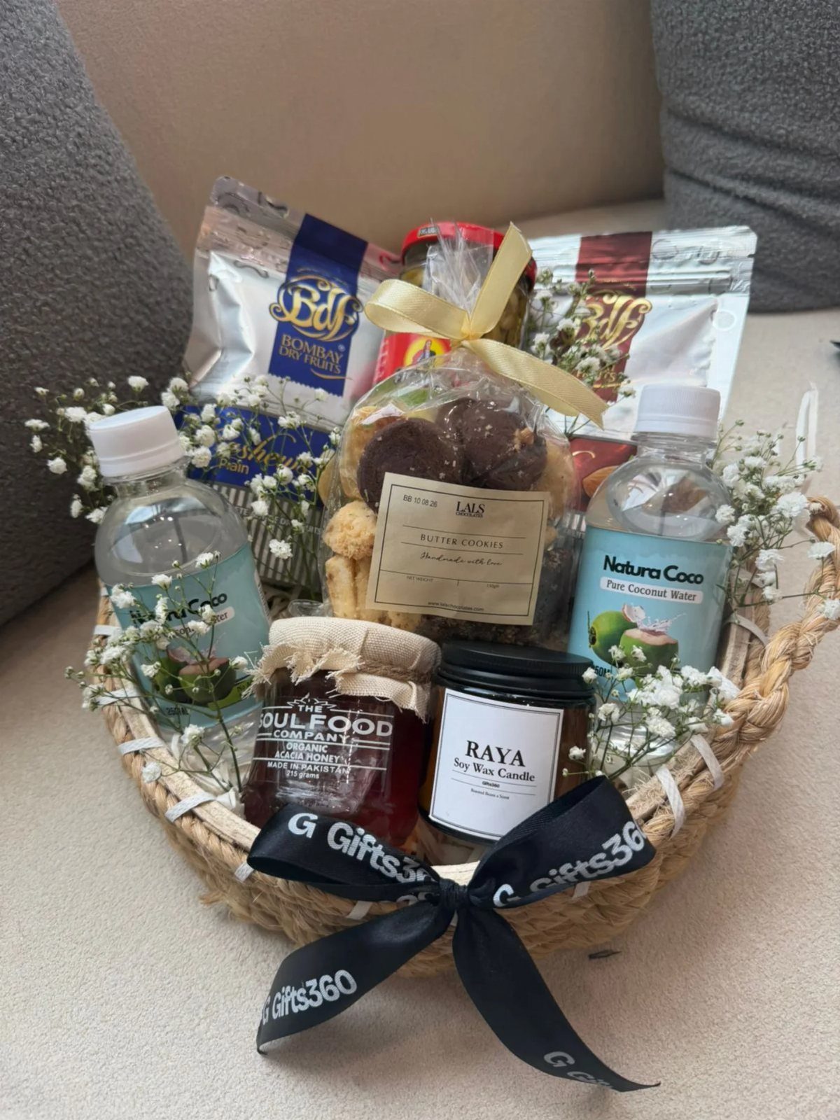 Wicker gift basket containing snacks, water, jam, and a candle with white flowers.