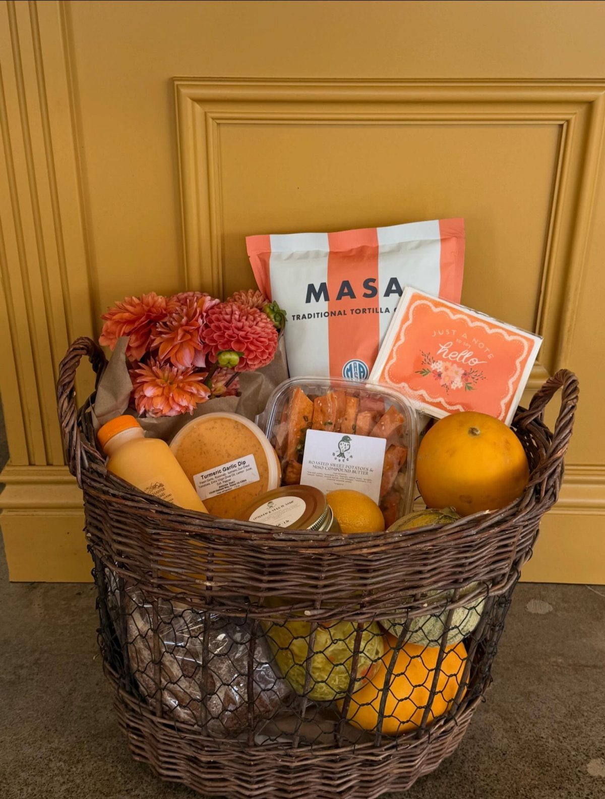 Woven gift basket containing fresh oranges, tortillas, and a bouquet of seasonal dahlias.