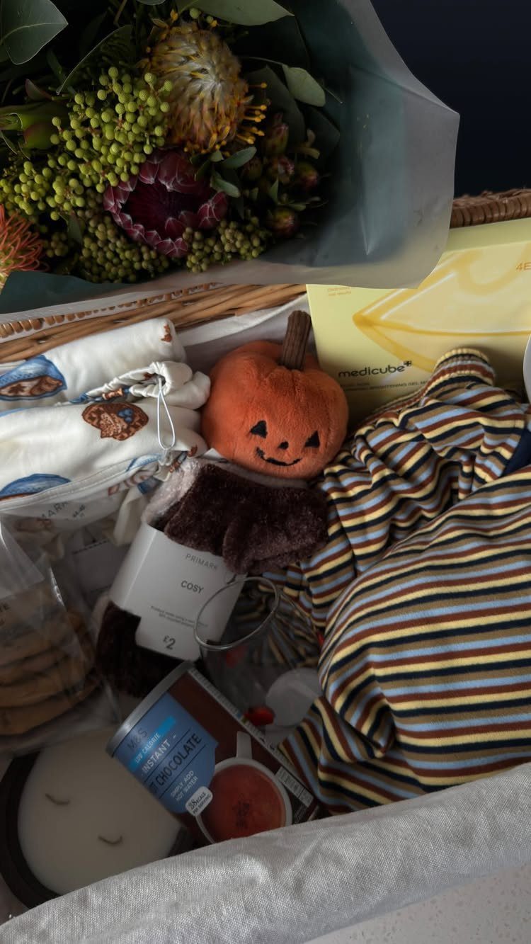 Wicker gift basket containing a plush pumpkin, striped socks, and a floral bouquet.
