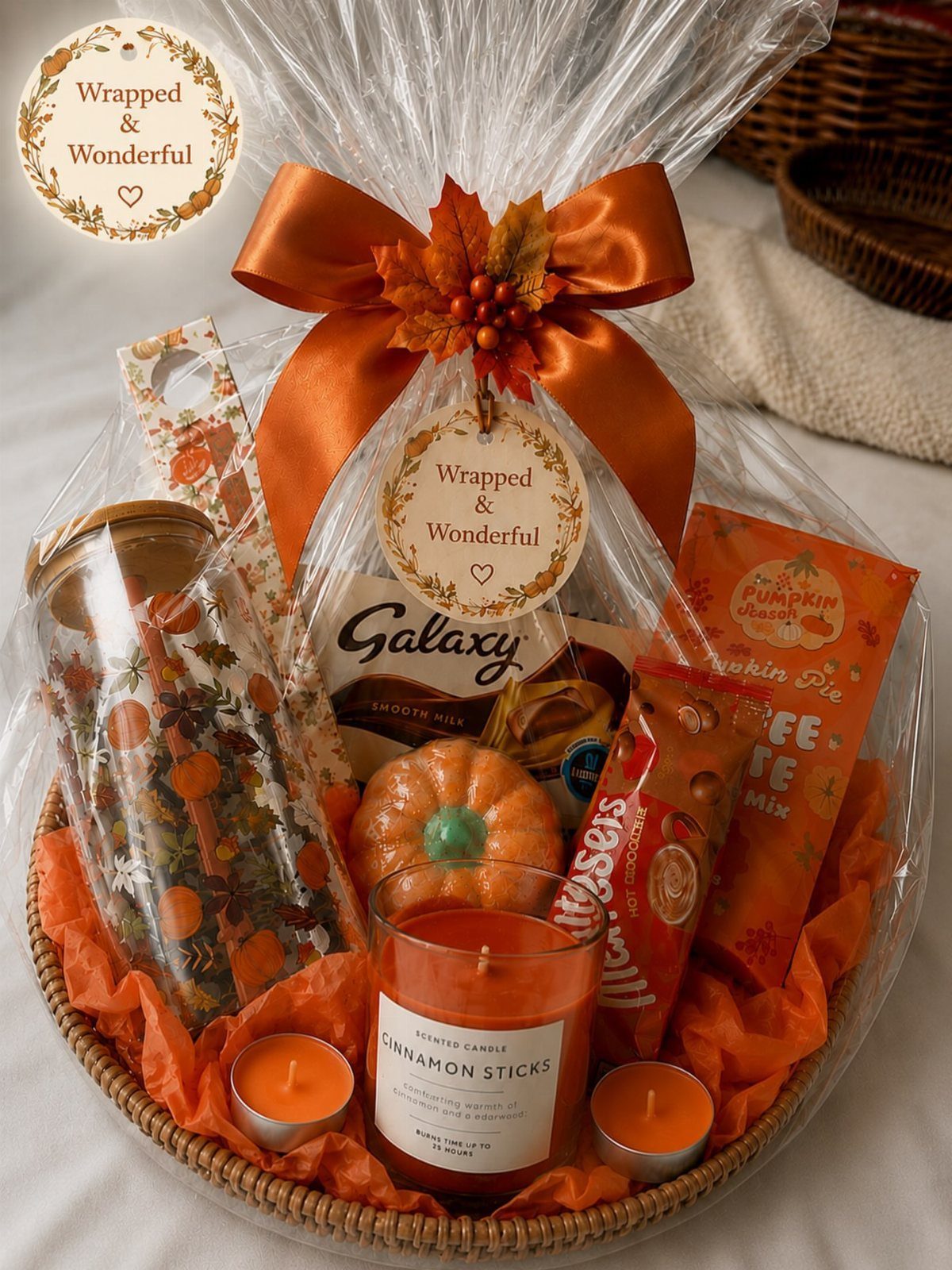 Wicker gift basket with orange bow containing pumpkin candles and autumn themed snacks.