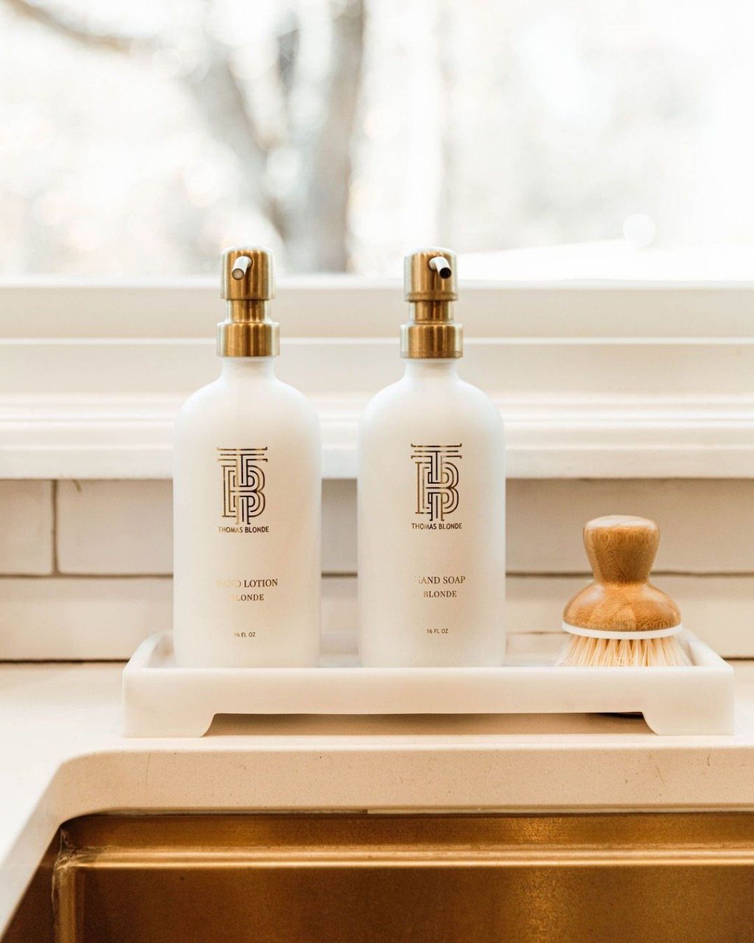Two white soap and lotion bottles with gold pumps on a marble tray.