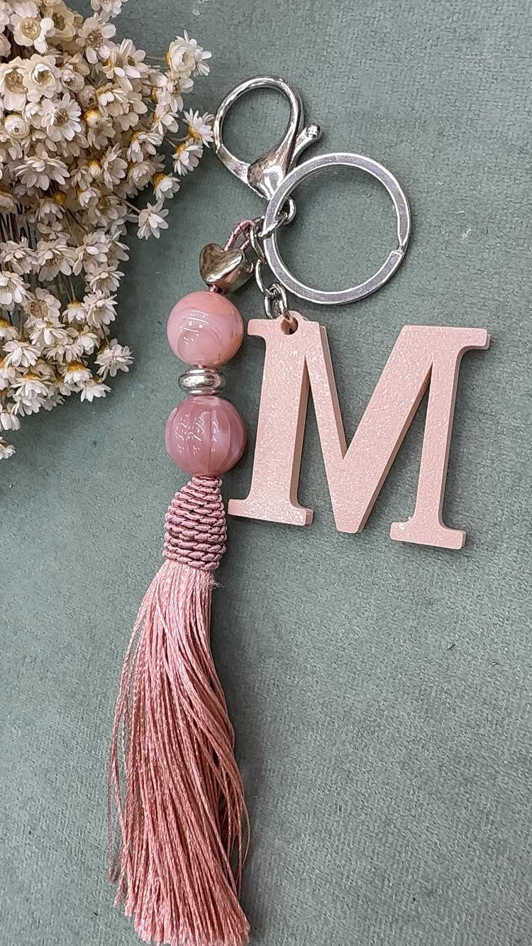 Pink keychain with letter M, beads, and tassel next to dried flowers.