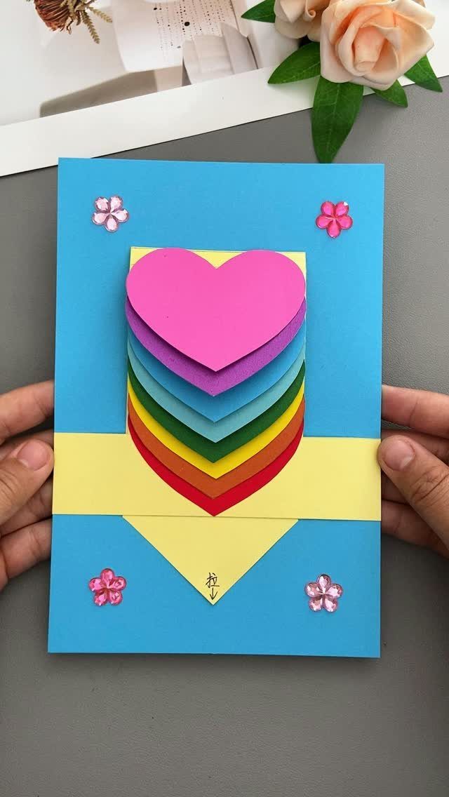 Handmade blue card with stacked rainbow paper hearts and small pink flower stickers.