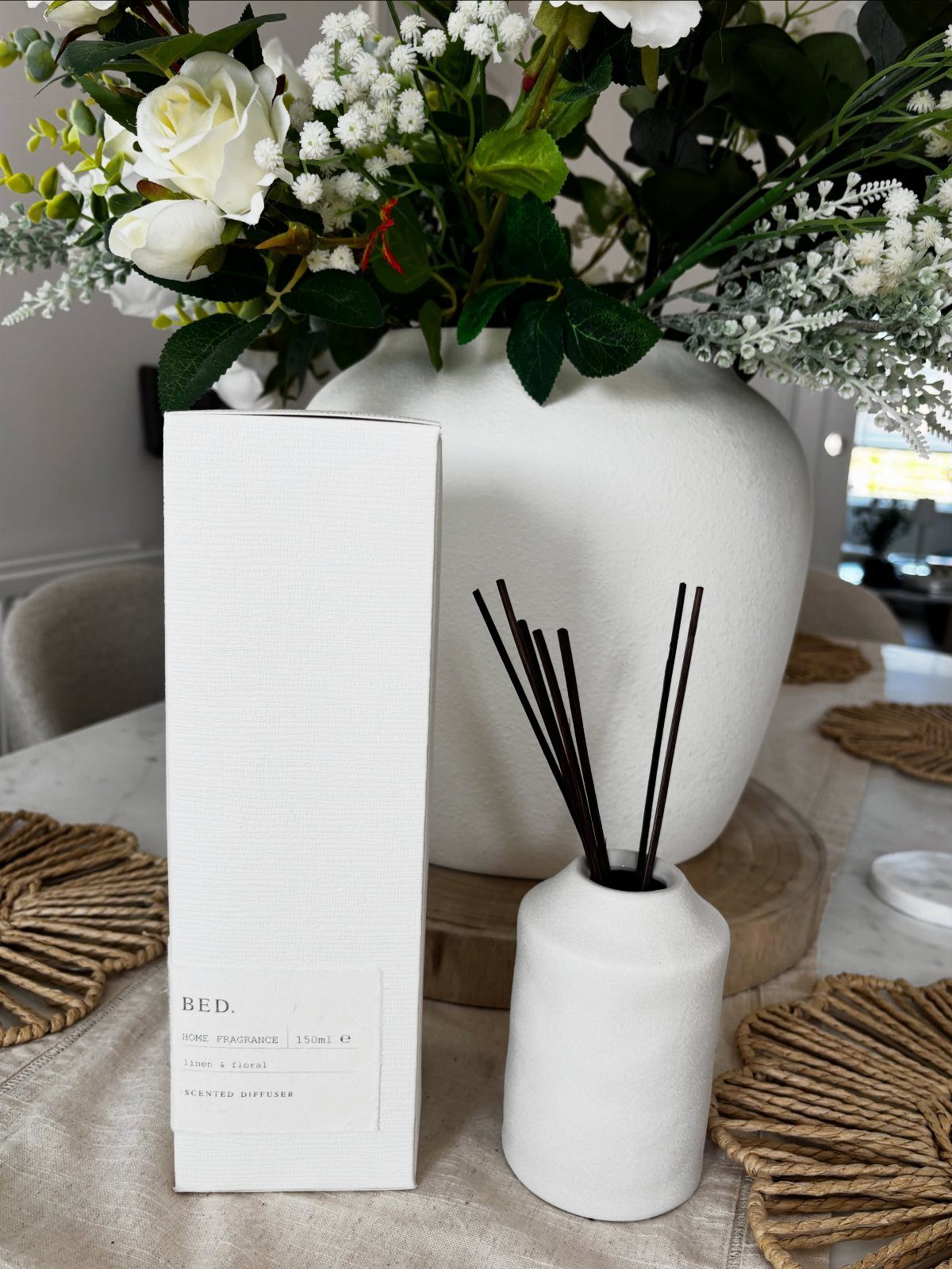 5 Housewarming Gift Ideas for Someone Who Has Everything 4 White ceramic reed diffuser with black sticks and packaging next to white roses.
