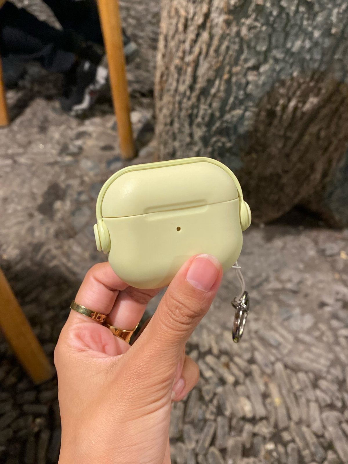 Light green earbud case held by a hand against a cobblestone background.